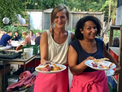 Zwei Frauen halten Teller mit Essen in die Kamera mit Festbänken im Hintergrund